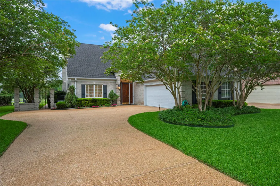 Exterior of a light gray brick house with a dark roof, a tan driveway, and green trees.