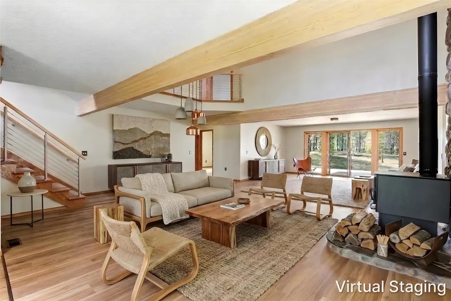 Bright living room with wood floors, beams, and a stone fireplace. A beige sofa and chairs sit on a jute rug. Sliding doors lead to a wooded view.