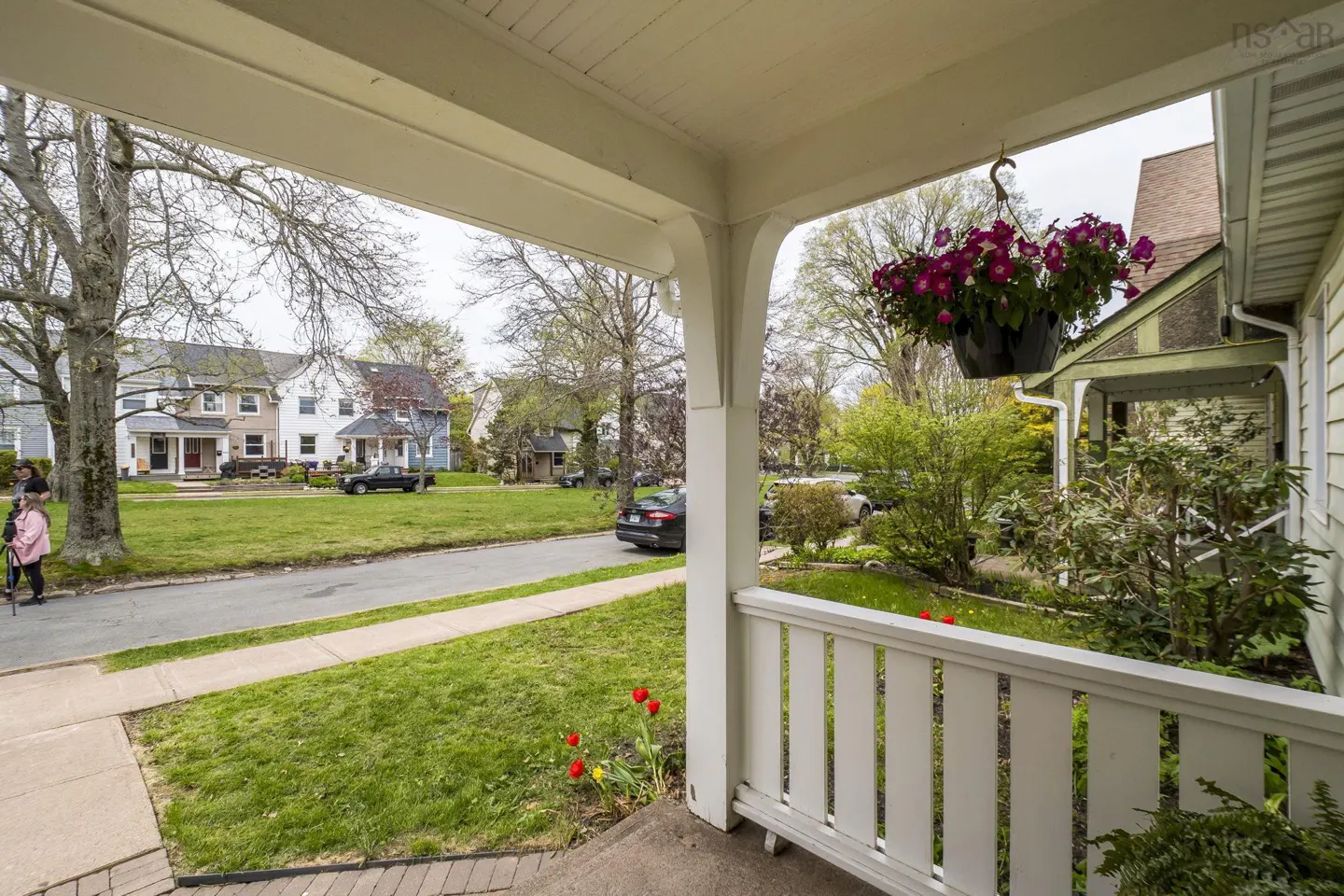 View from a porch with white railings and a hanging flower basket, overlooking a green lawn and street with houses.