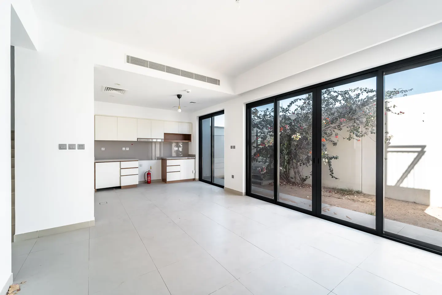 Bright, empty room with white walls, tile floor, and kitchen cabinets. Black-framed sliding glass doors lead to a small yard with a flowering bush.