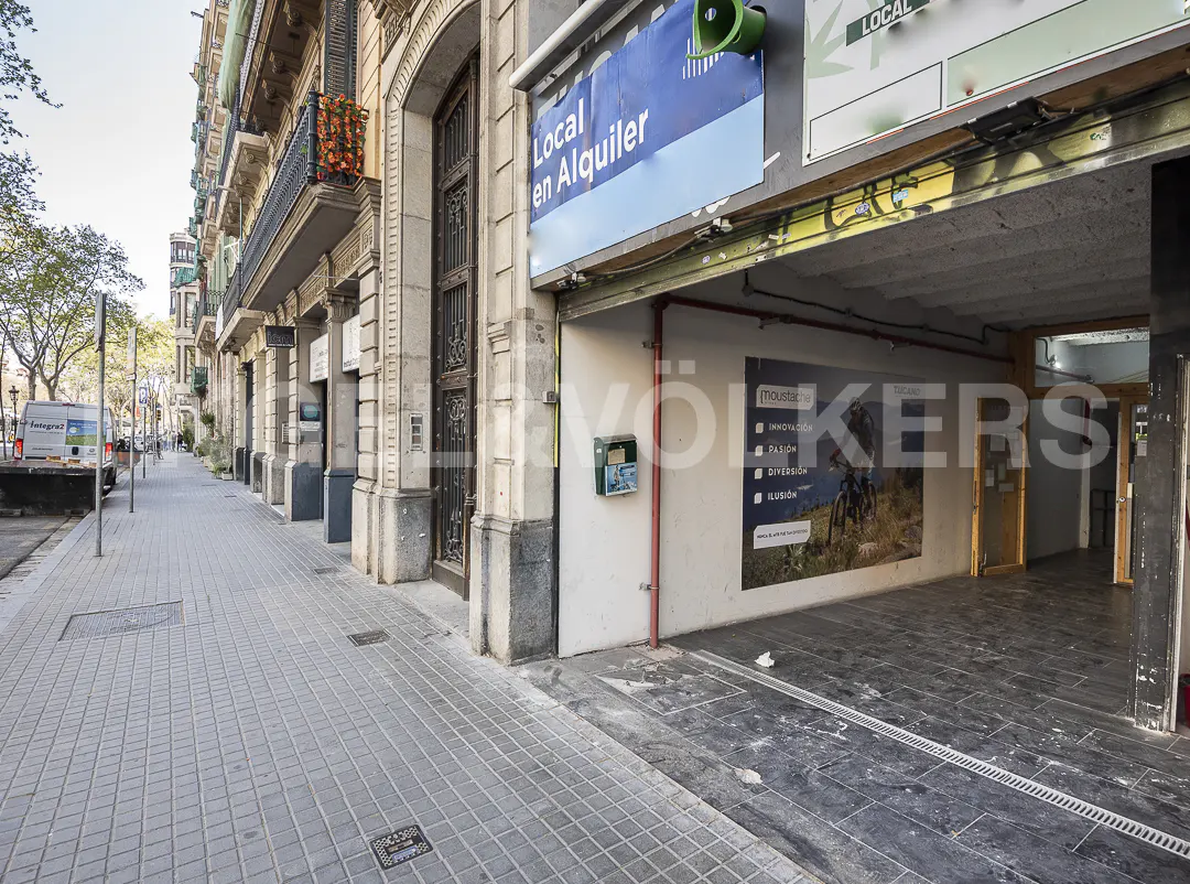 Street view of a commercial space for rent with a blue "Local en Alquiler" sign. The building has stone columns and balconies with flowers.