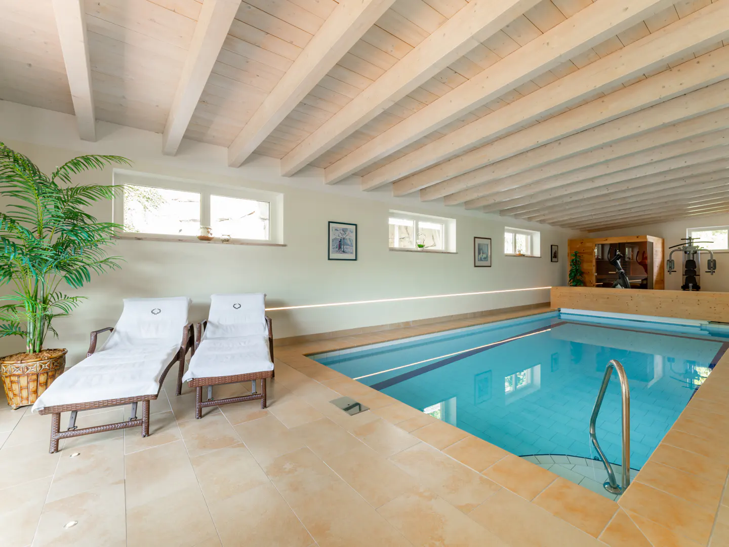 Indoor pool area with blue water, beige tile floor, white ceiling beams, two lounge chairs, and exercise equipment in the background.