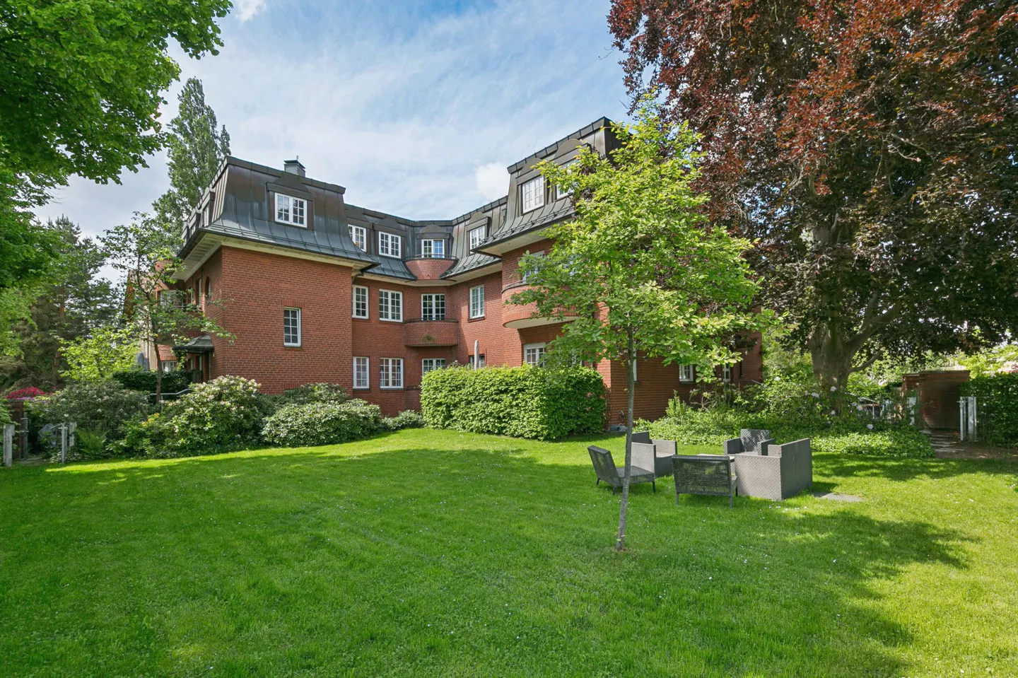 Brick apartment building with a dark roof, surrounded by green lawn, trees, and an outdoor seating area.