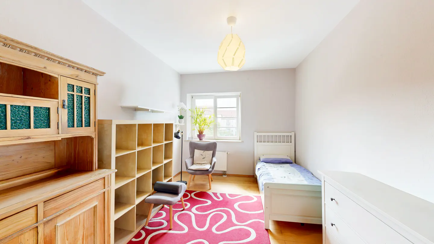 Bright bedroom with a single bed, wooden dresser, and a red patterned rug. A window provides natural light.