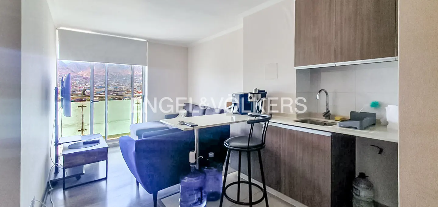 Studio apartment with a blue sofa, a white table, a black stool, and a kitchen with brown cabinets. A window shows a city view.