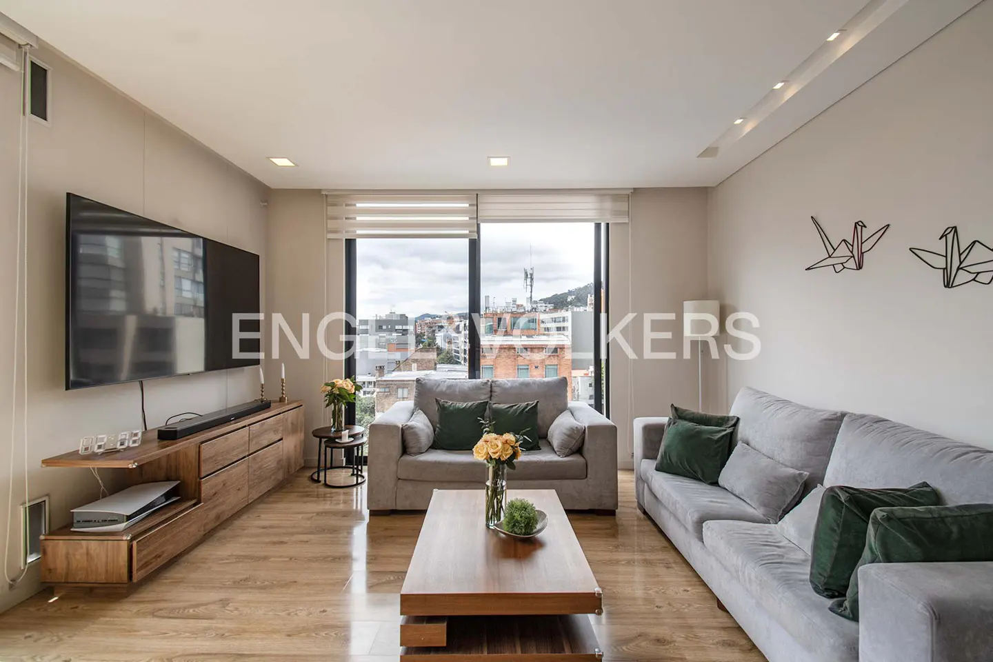Living room with gray sofas, wood floors, and a large window with a city view. A wood TV stand holds a large screen TV.