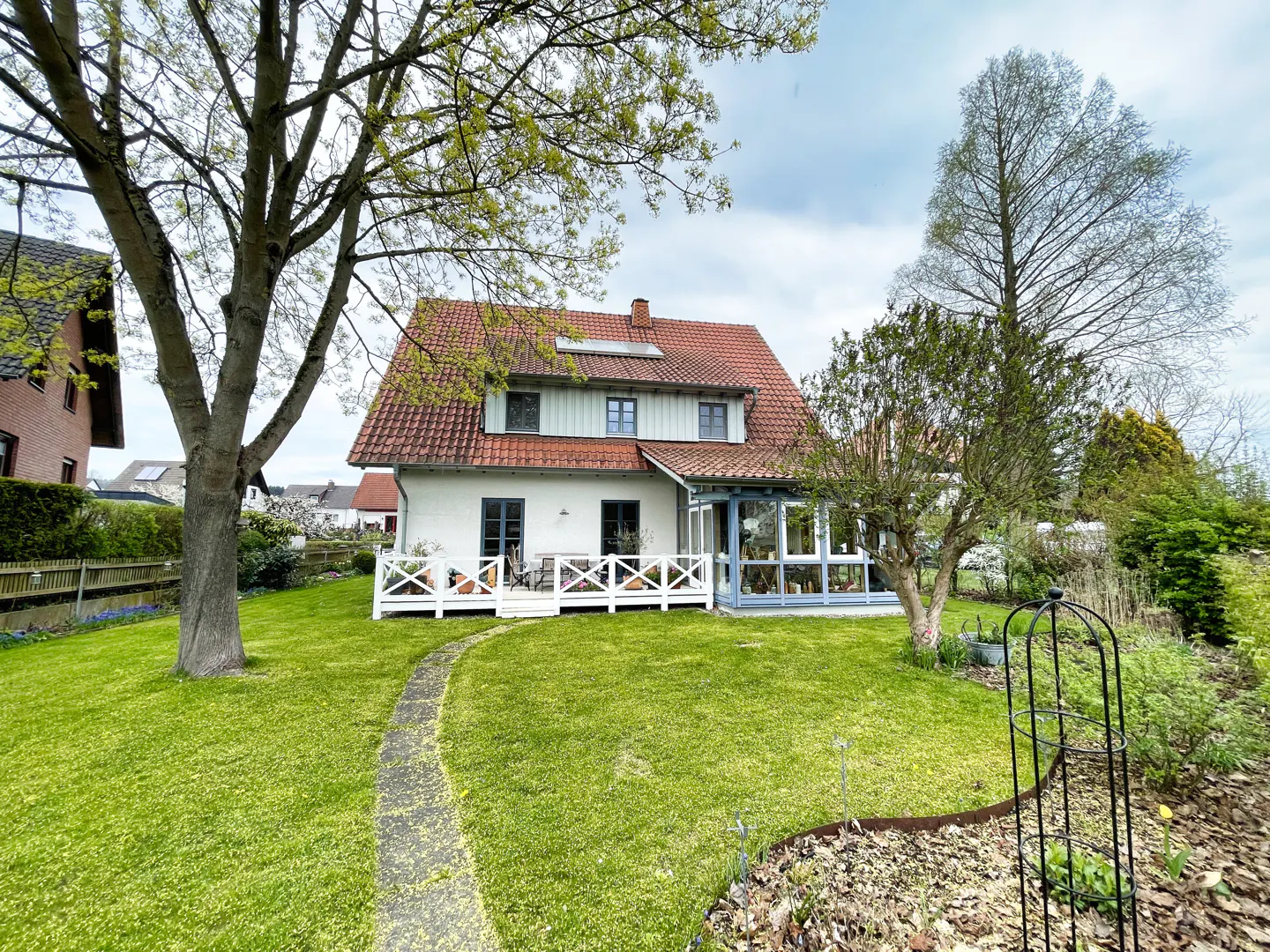 Two-story white house with a red tile roof, white deck, and glass sunroom, surrounded by a green lawn and trees.