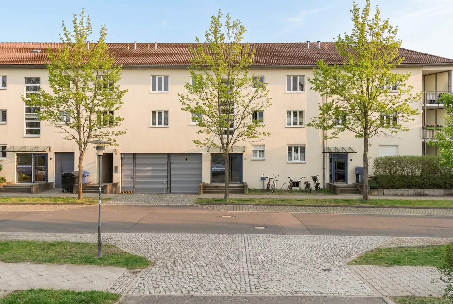 Three-story apartment building with a red tile roof and cream-colored walls, framed by young green trees.