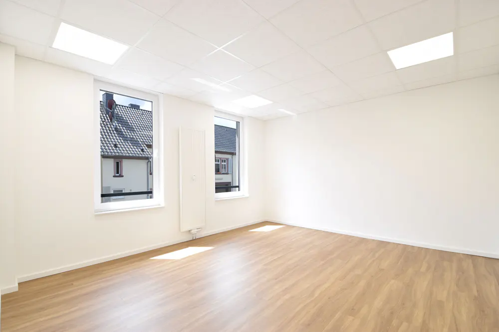 Empty room with white walls, wood-look flooring, recessed ceiling lights, and two windows overlooking a residential street.