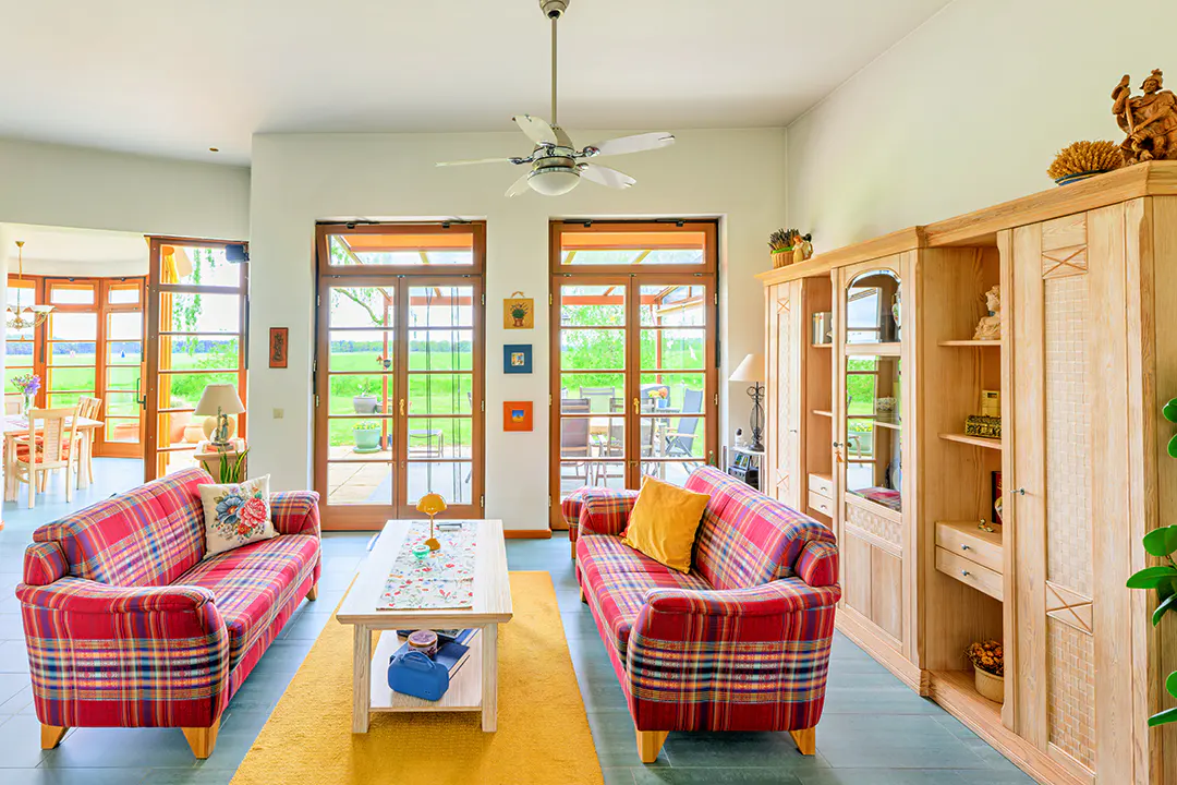 Bright living room with two red plaid sofas, a light wood cabinet, and French doors opening to a green lawn.