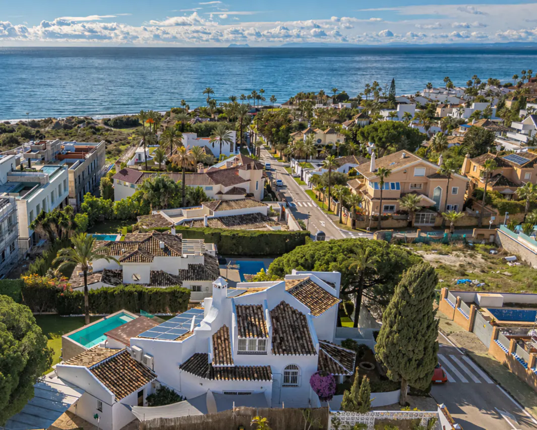 Aerial view of a coastal neighborhood with white houses, terracotta roofs, and the blue ocean in the background.