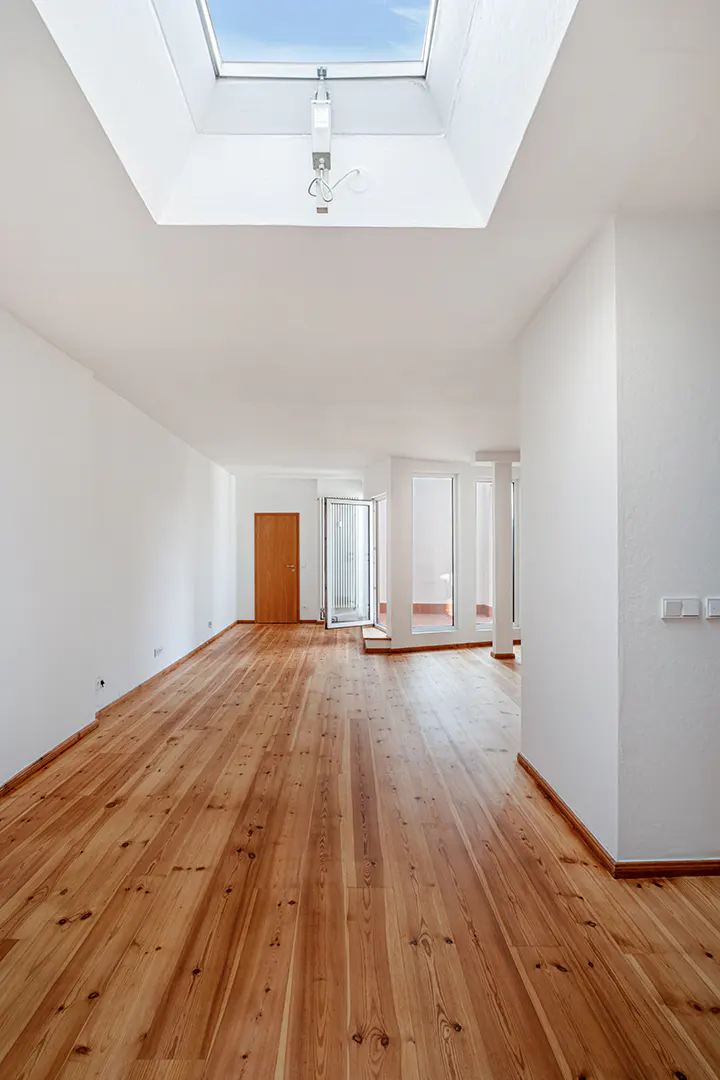 Bright, empty room with wood floors, white walls, and a skylight. A brown door and windows are visible in the background.