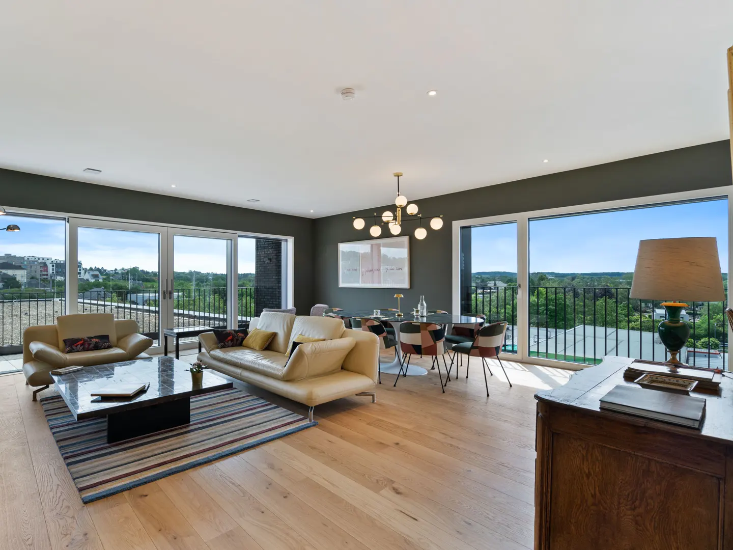 Bright, modern living room with light wood floors, cream sofas, and a dining area. Large windows offer a view of the outside.