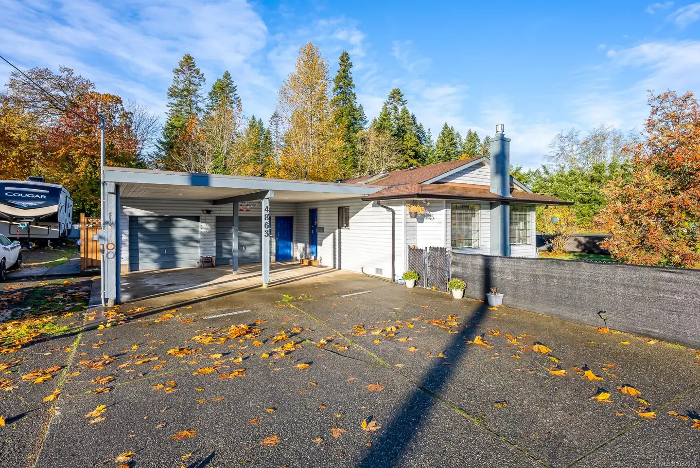 A single-story white house with a blue door and a gray carport on a sunny day with autumn leaves.
