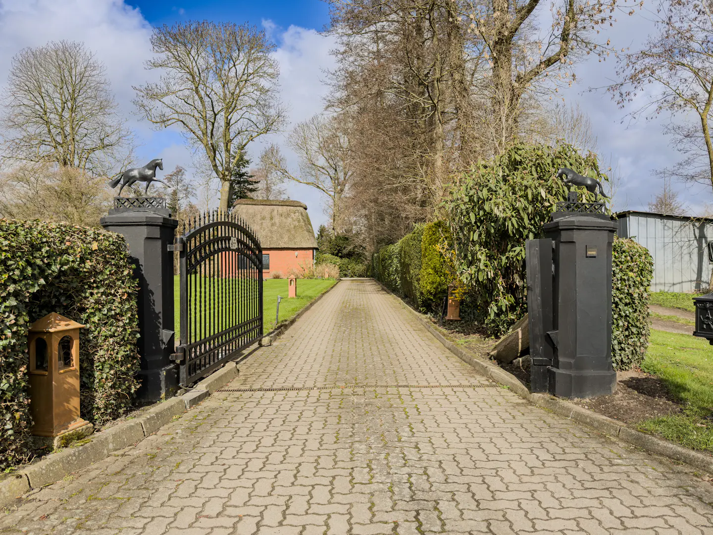 Gated driveway leads to a red house with a thatched roof. Black iron gates are topped with horse statues. Green hedges line the drive.
