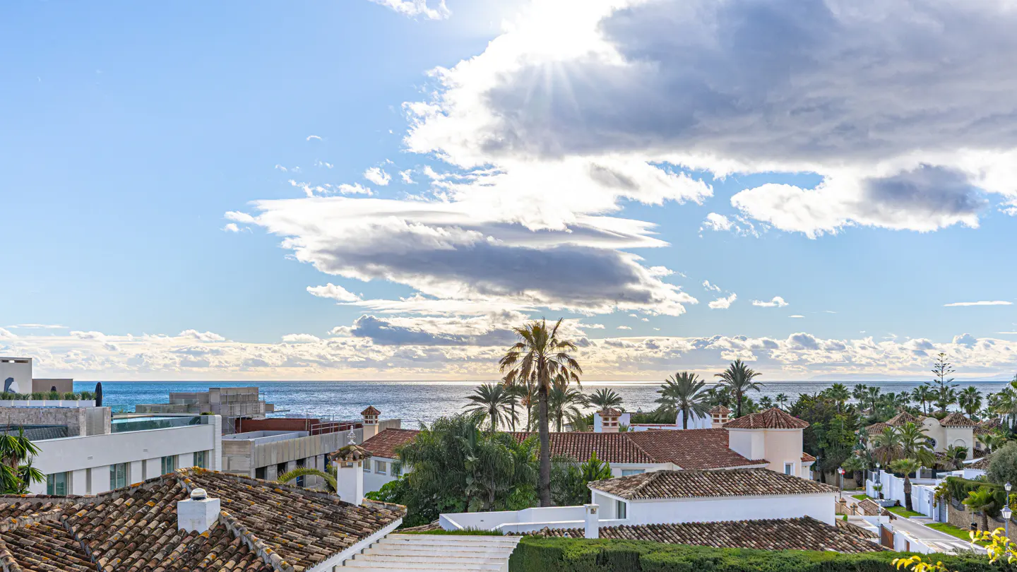 View of a coastal town with red-tiled roofs, palm trees, and the ocean under a blue sky with white clouds.