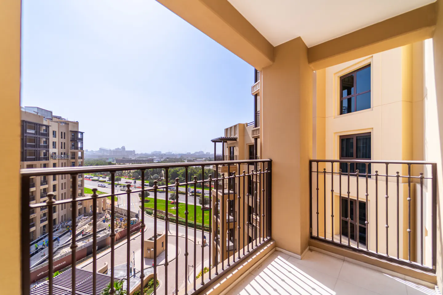 View from a beige balcony with black metal railings overlooking a street with trees and buildings.