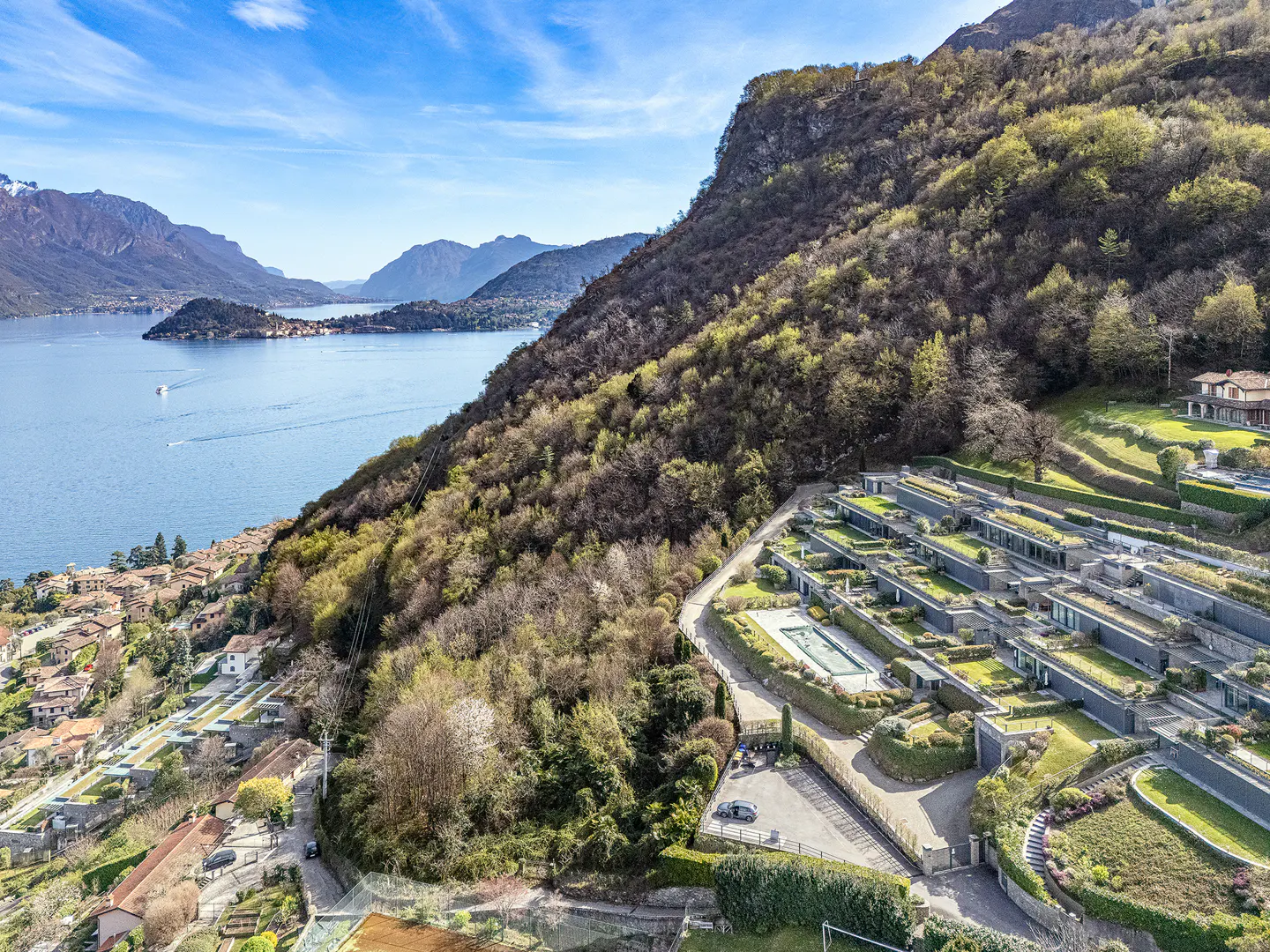 Lake Como view with modern hillside villas. Blue water, mountains, and green roofs under a bright sky.