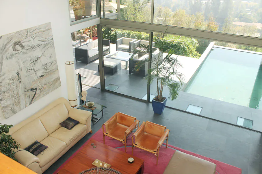 Aerial view of a modern living room with a cream sofa, leather chairs, and a pink rug. Outside, a patio with furniture and a pool are visible through large windows.