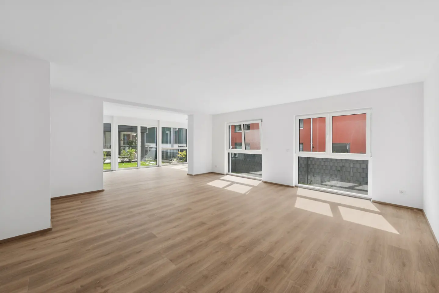 Bright, empty room with light wood floors, white walls, and large windows. Outside, greenery and a red building are visible.