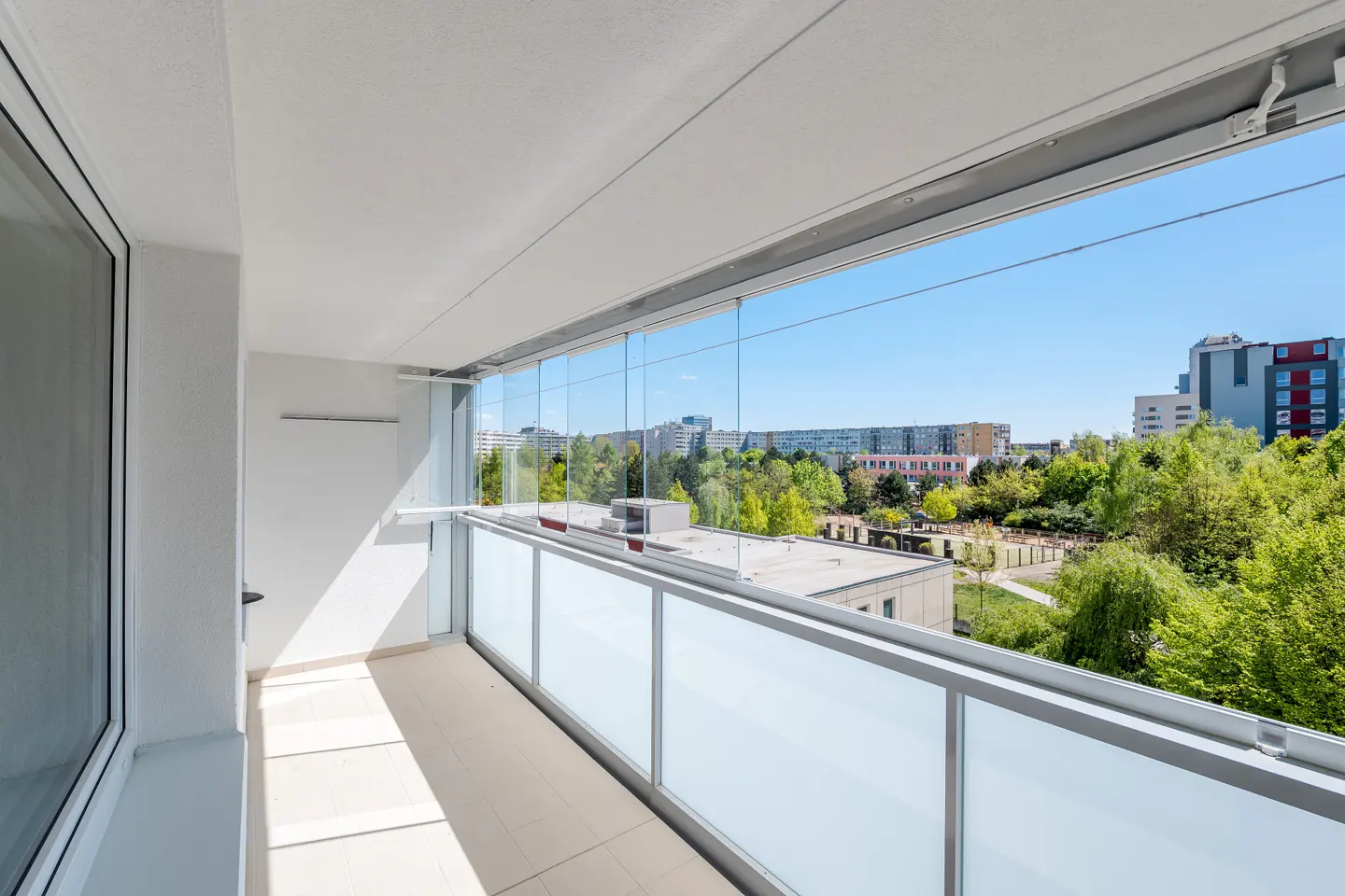 Modern balcony with glass railings and sliding panels, overlooking a sunny urban landscape with trees and buildings.
