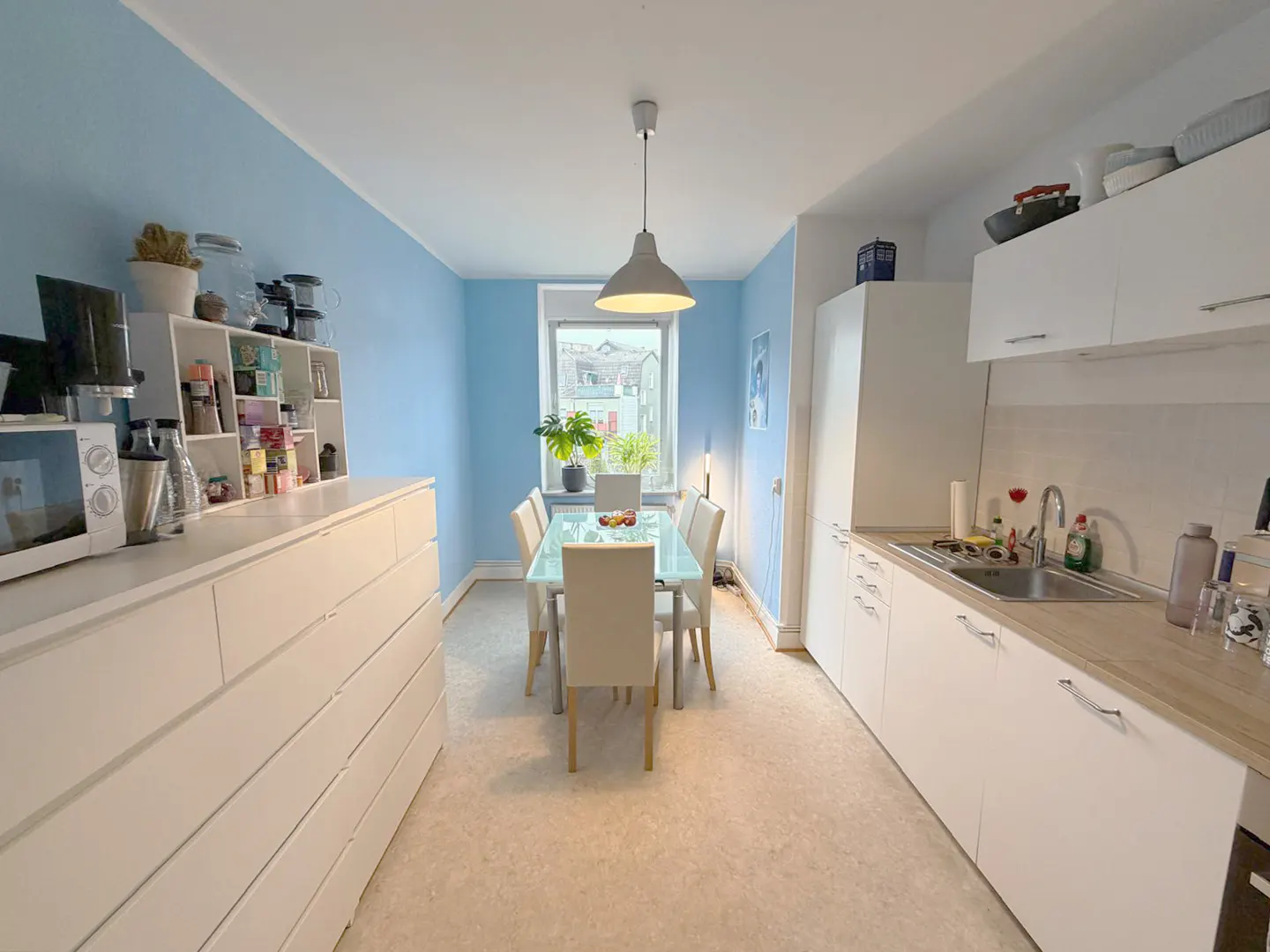 A bright kitchen with light blue walls, white cabinets, and a glass dining table with six chairs. A window is visible in the background.