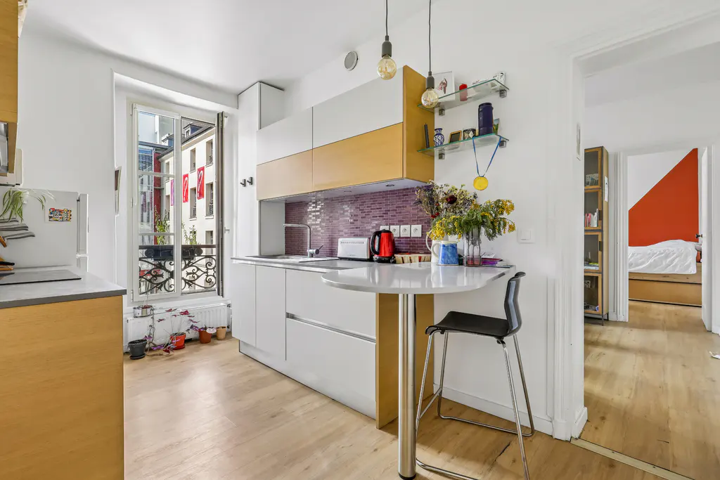 Bright kitchen with white cabinets, wood floors, and a view of buildings with red flags. A round table with a black stool sits in the foreground.