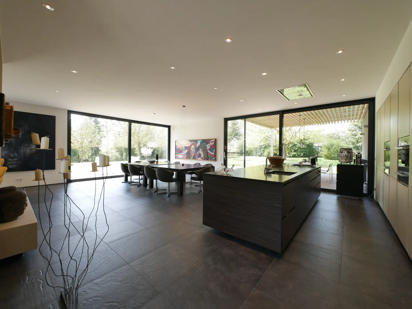 Open-concept kitchen and dining area with dark tile floors and large windows. A dark island sits in the kitchen, and a dining table with chairs is visible.