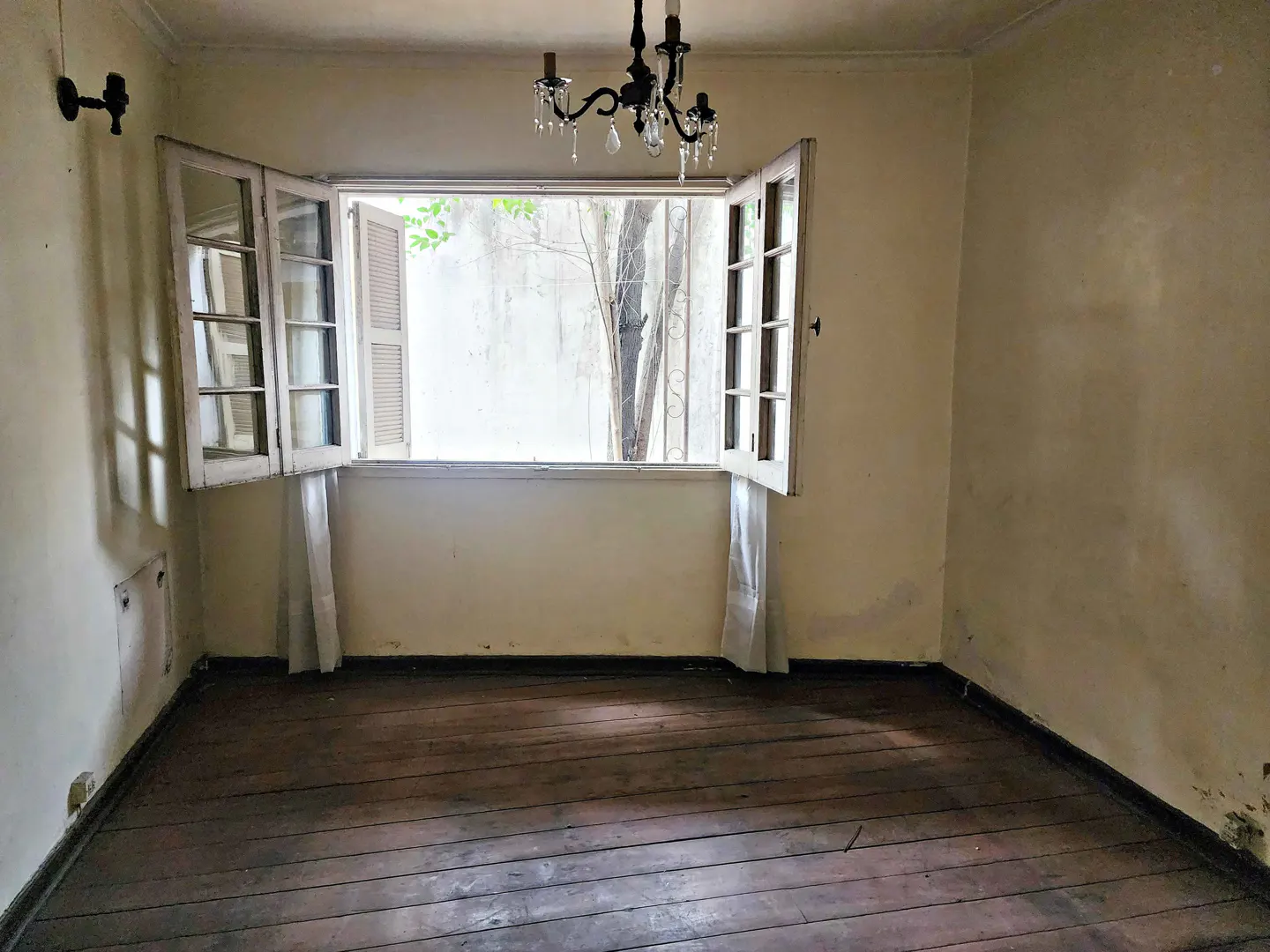 Empty room with wood floors, cream walls, and open white windows. A black chandelier hangs from the ceiling.