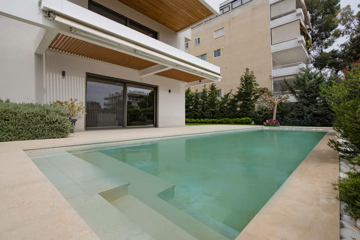 A modern white house with a turquoise swimming pool and stone steps leading into the water. Green trees and a building are in the background.