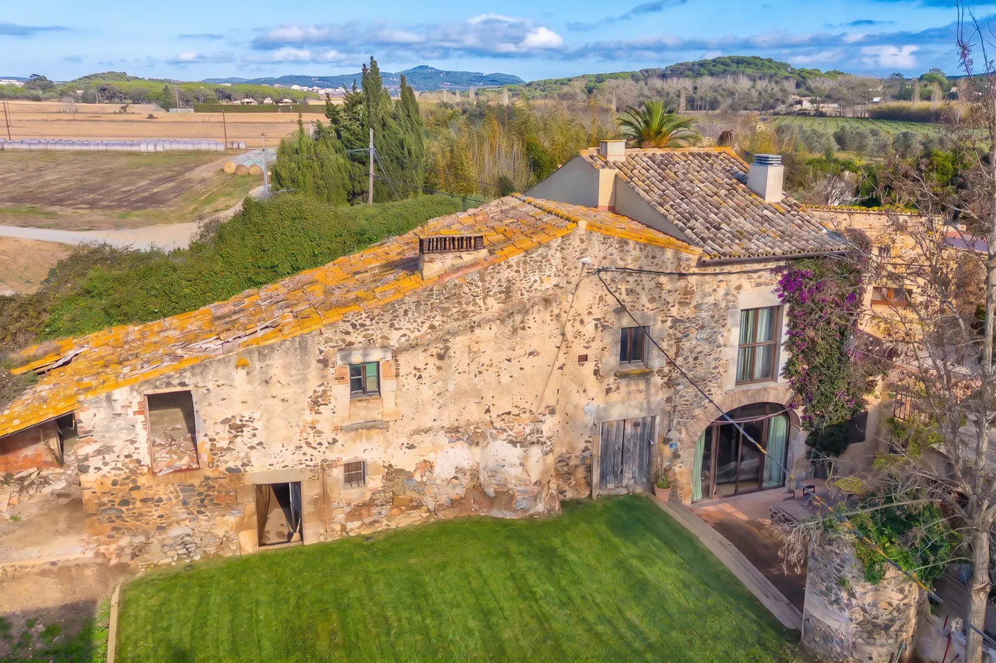 Aerial view of a stone farmhouse with a mossy tile roof and green lawn in a rural setting.