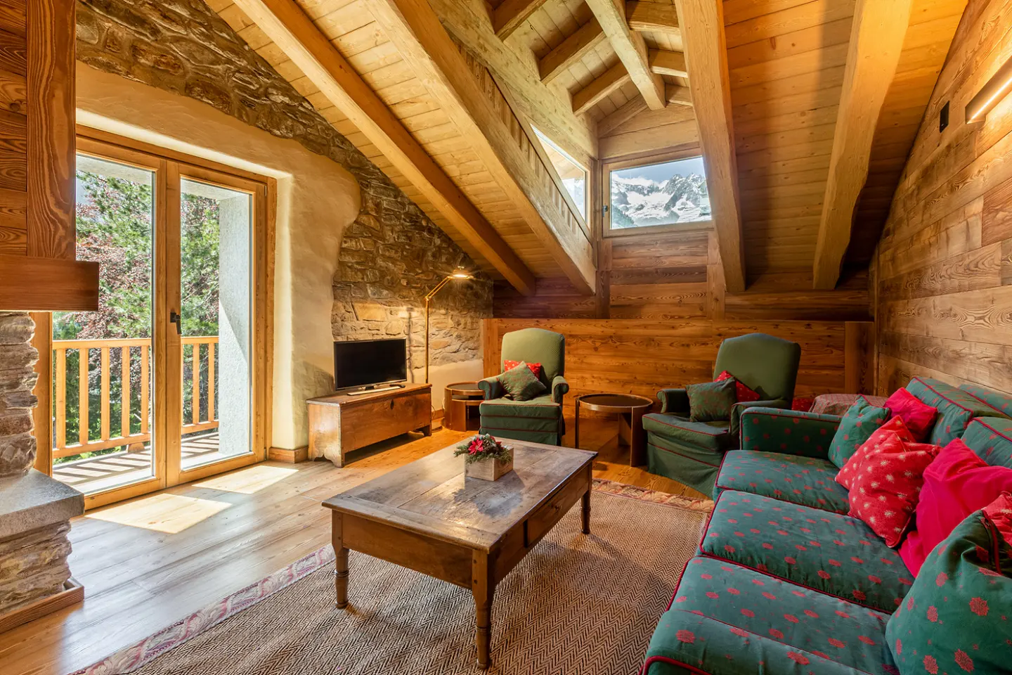 Cozy rustic attic living room with wood-paneled walls, exposed beams, green armchairs, a sofa, and mountain views.