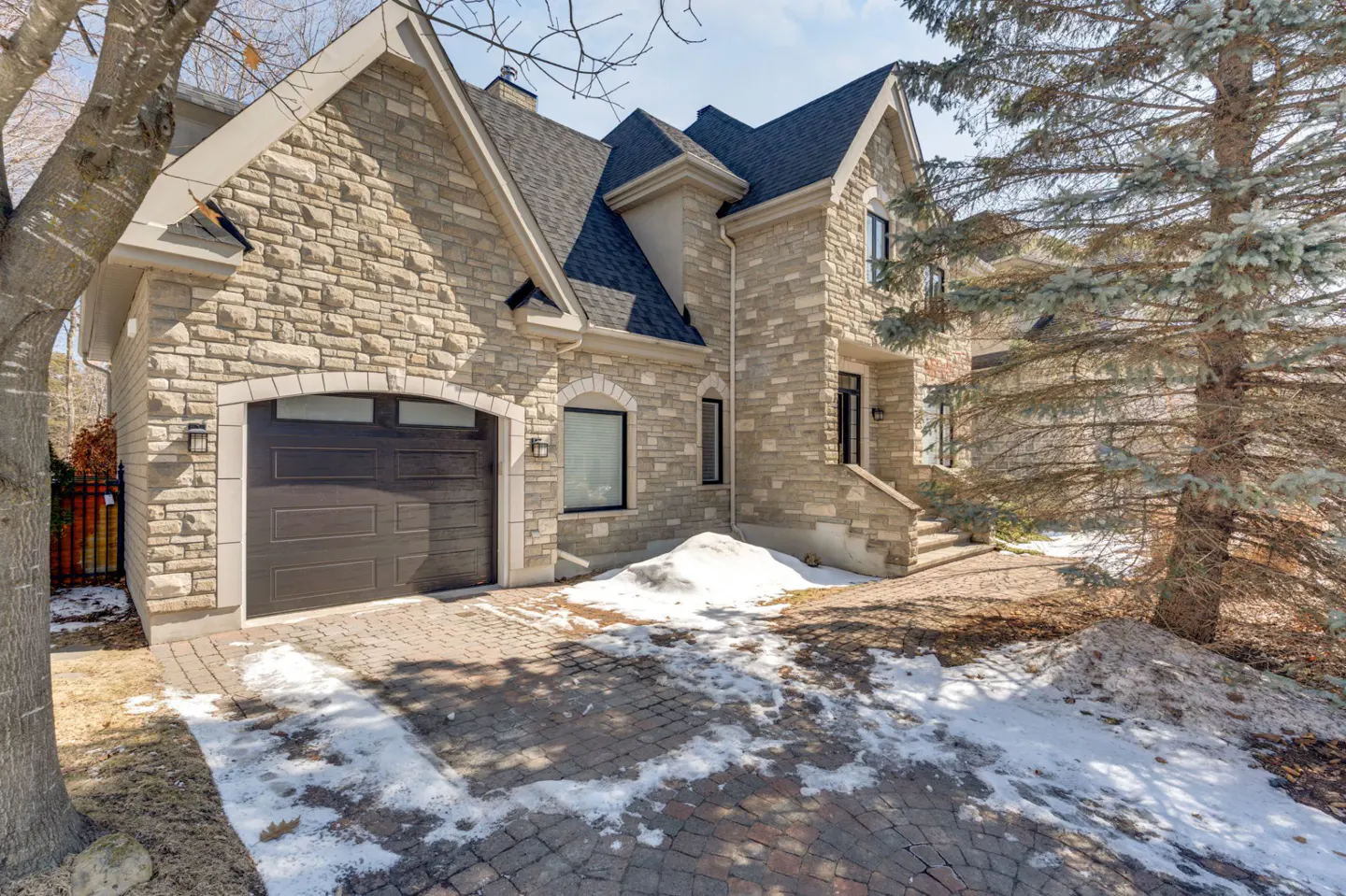 Stone house with a dark roof, brown garage door, and brick driveway with snow patches. A large pine tree stands to the right.