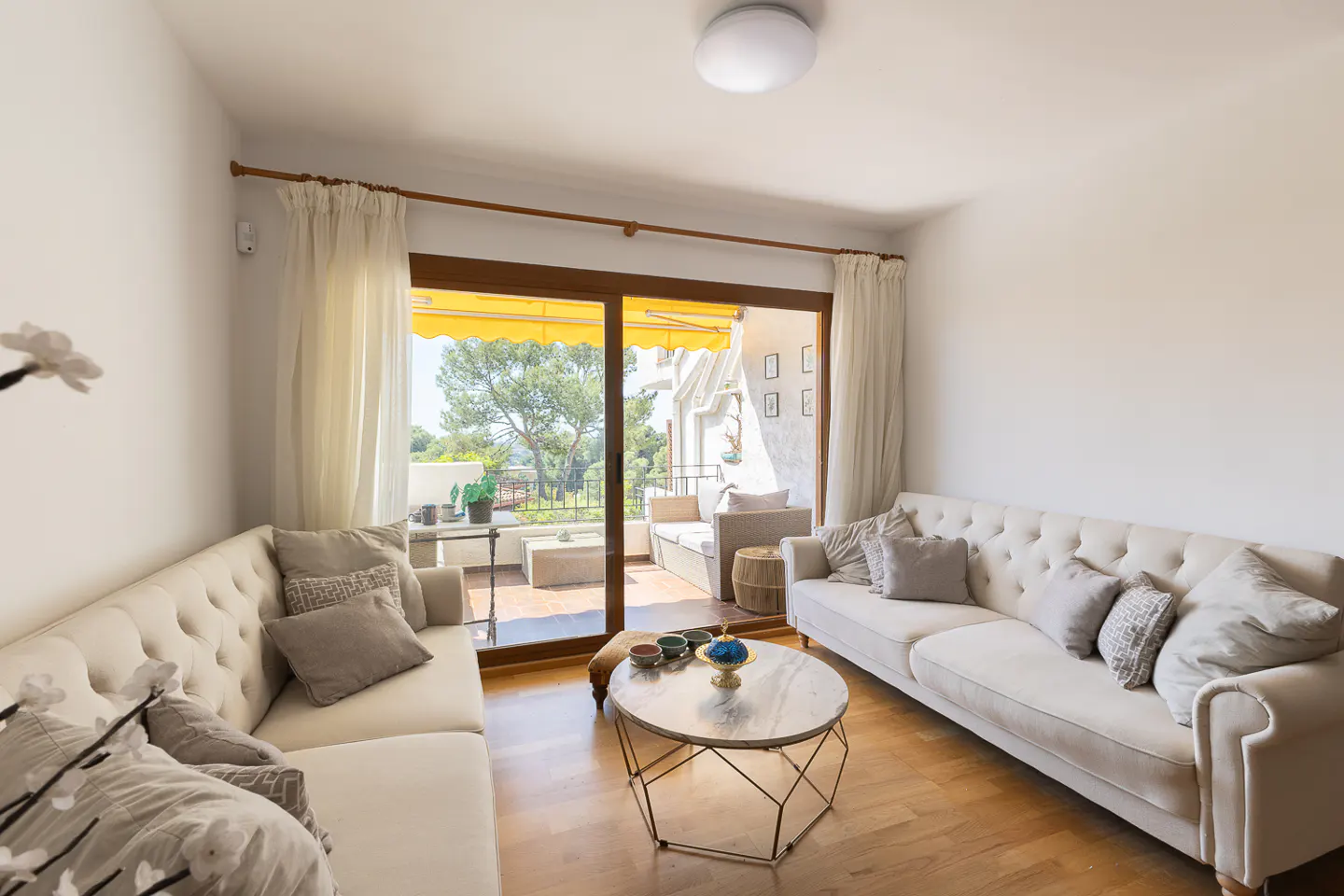 Bright living room with hardwood floors, white walls, and two sofas. Sliding glass doors lead to a balcony with a yellow awning and outdoor seating.