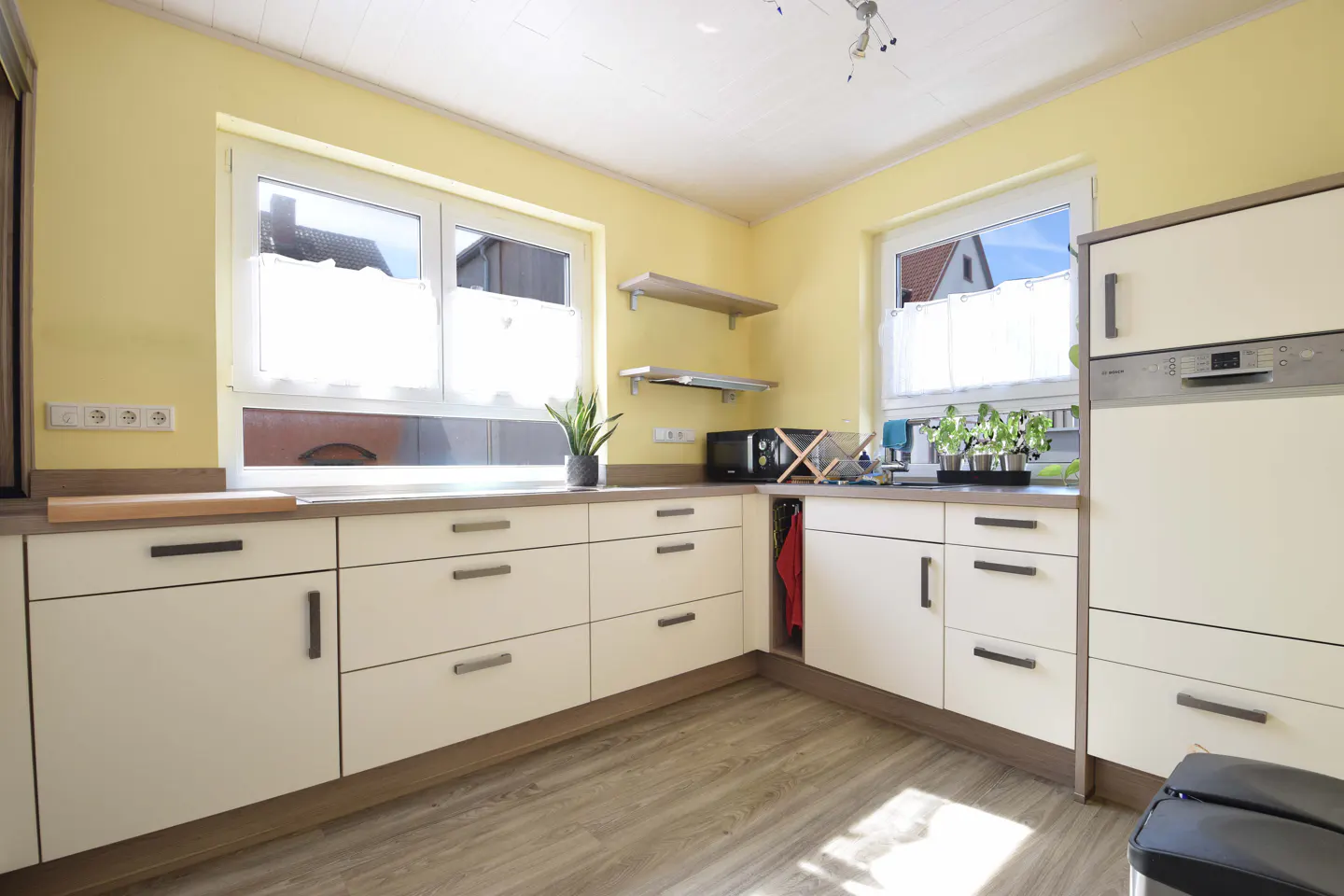 Bright kitchen with yellow walls, white cabinets, and wood-look flooring. Windows with sheer curtains let in natural light.