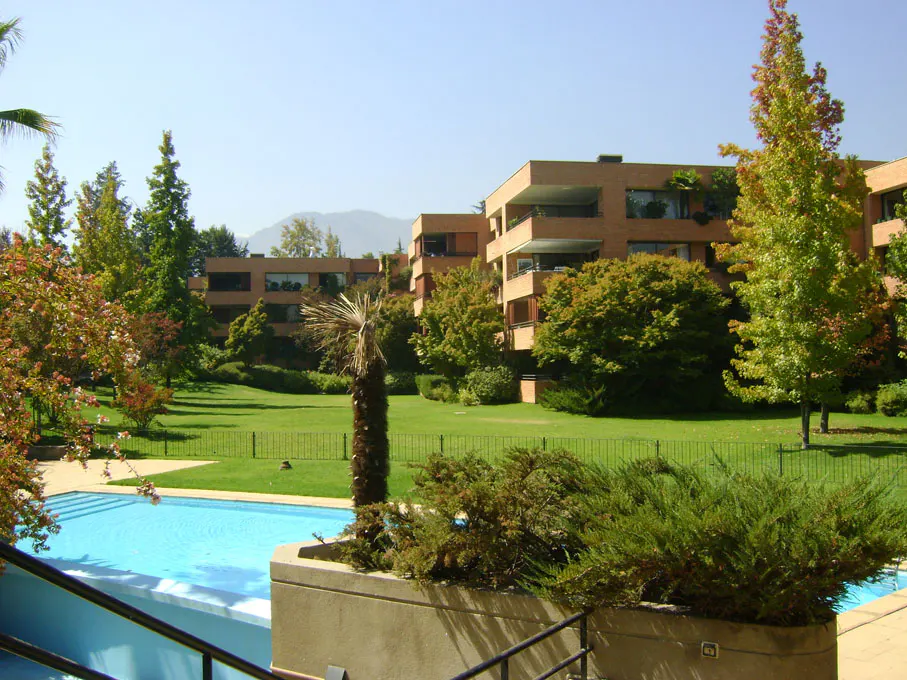 Exterior view of a modern apartment complex with a pool, green lawn, trees, and mountains in the background.