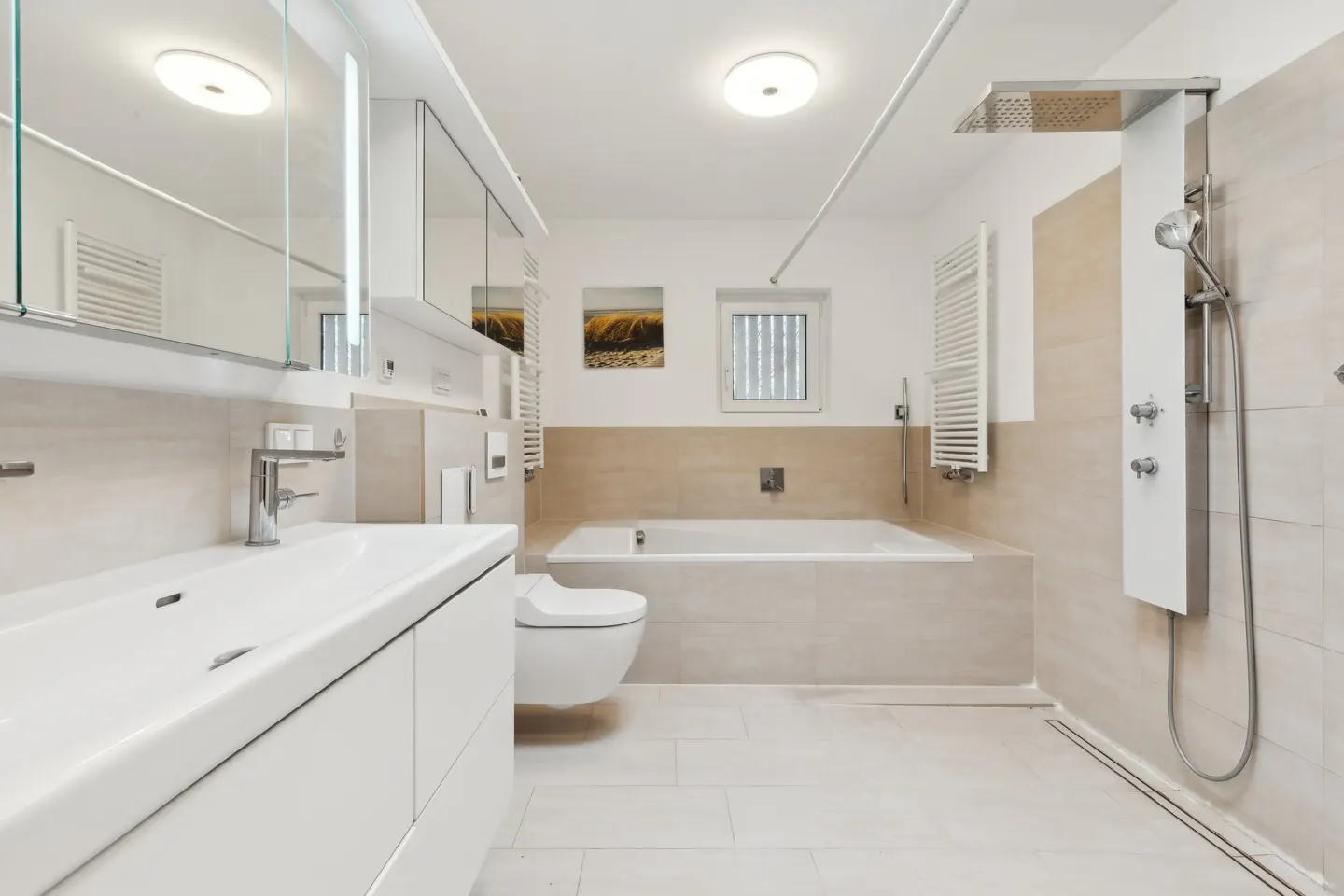 Bright bathroom with white vanity, toilet, and tub. Beige tile accents the walls. A modern shower panel stands to the right.