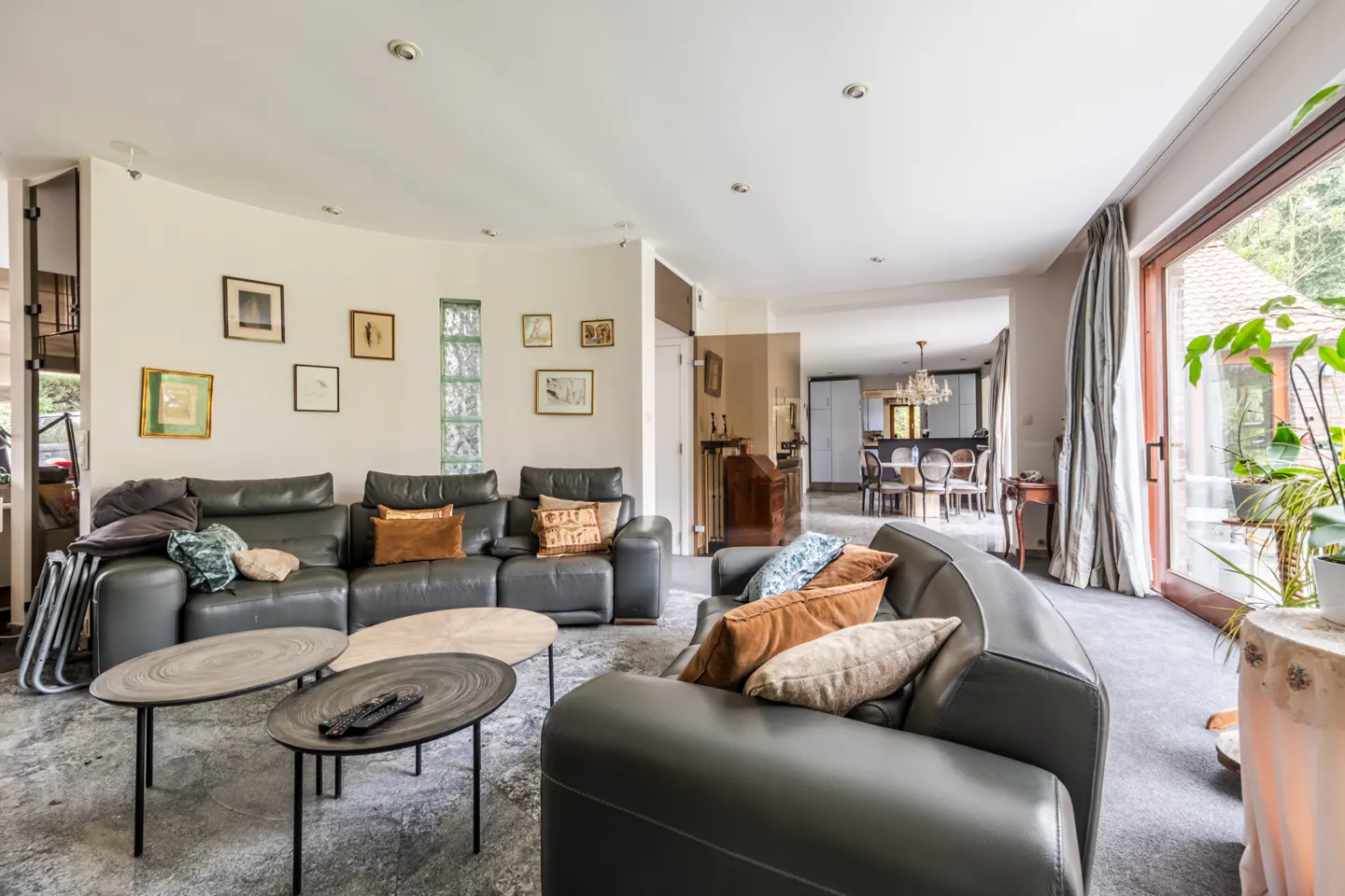 Living room with gray sofas, brown pillows, and oval tables. Art on the walls, and a view into the dining room. Large glass doors to the outside.