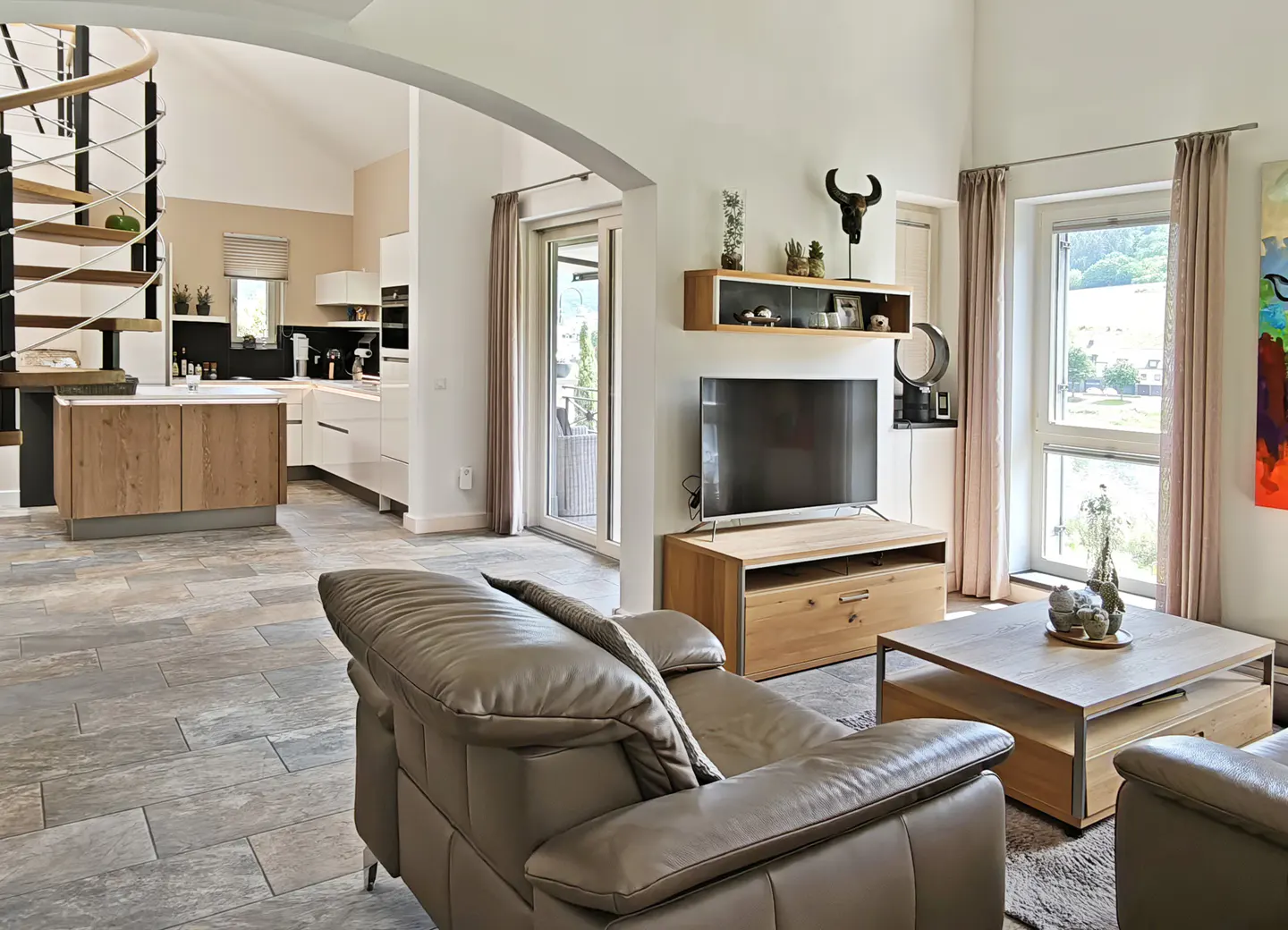 Open-plan living room with grey leather sofa, wooden coffee table, TV, and spiral staircase leading to a modern kitchen.