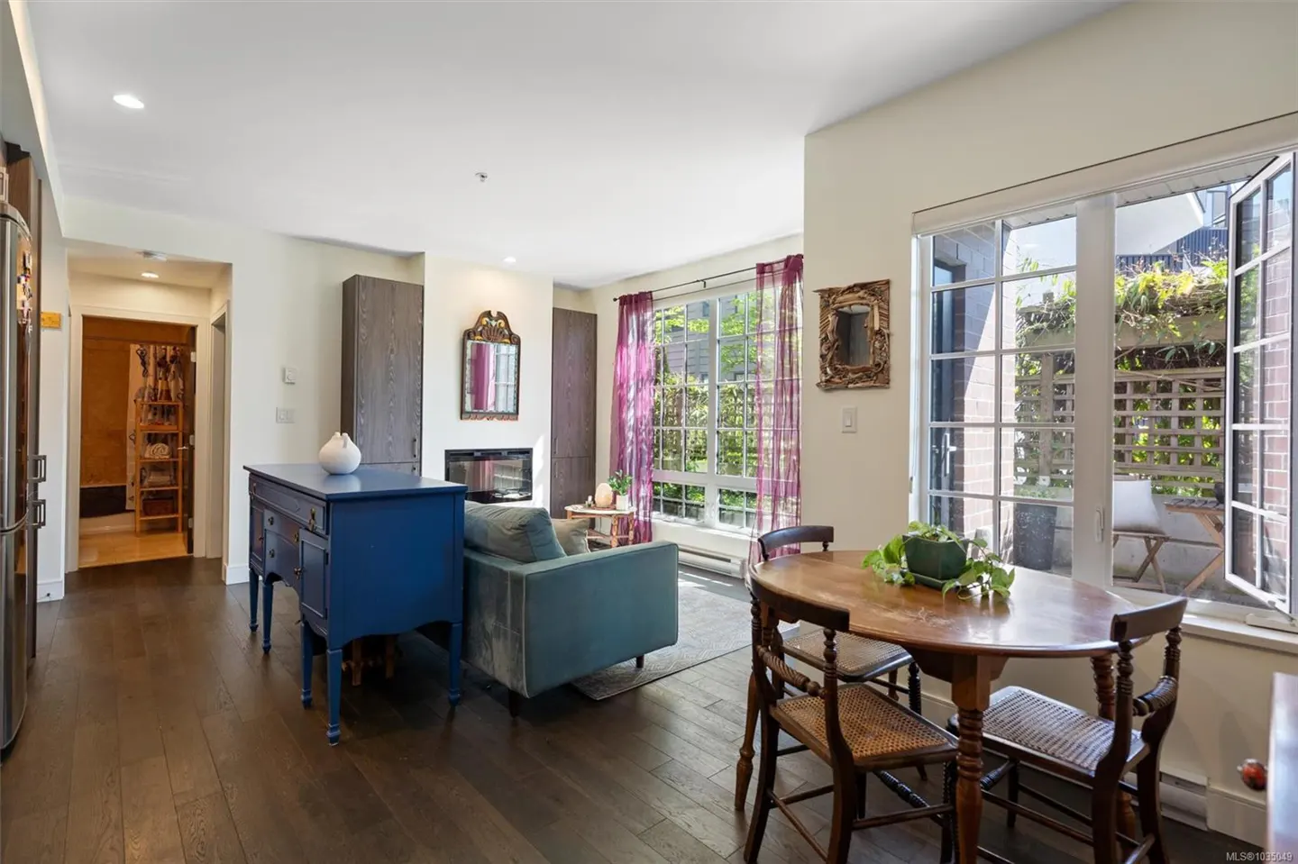Bright living space with dark wood floors, blue accent table, and dining set by a window overlooking a patio. A fireplace and teal sofa are visible.