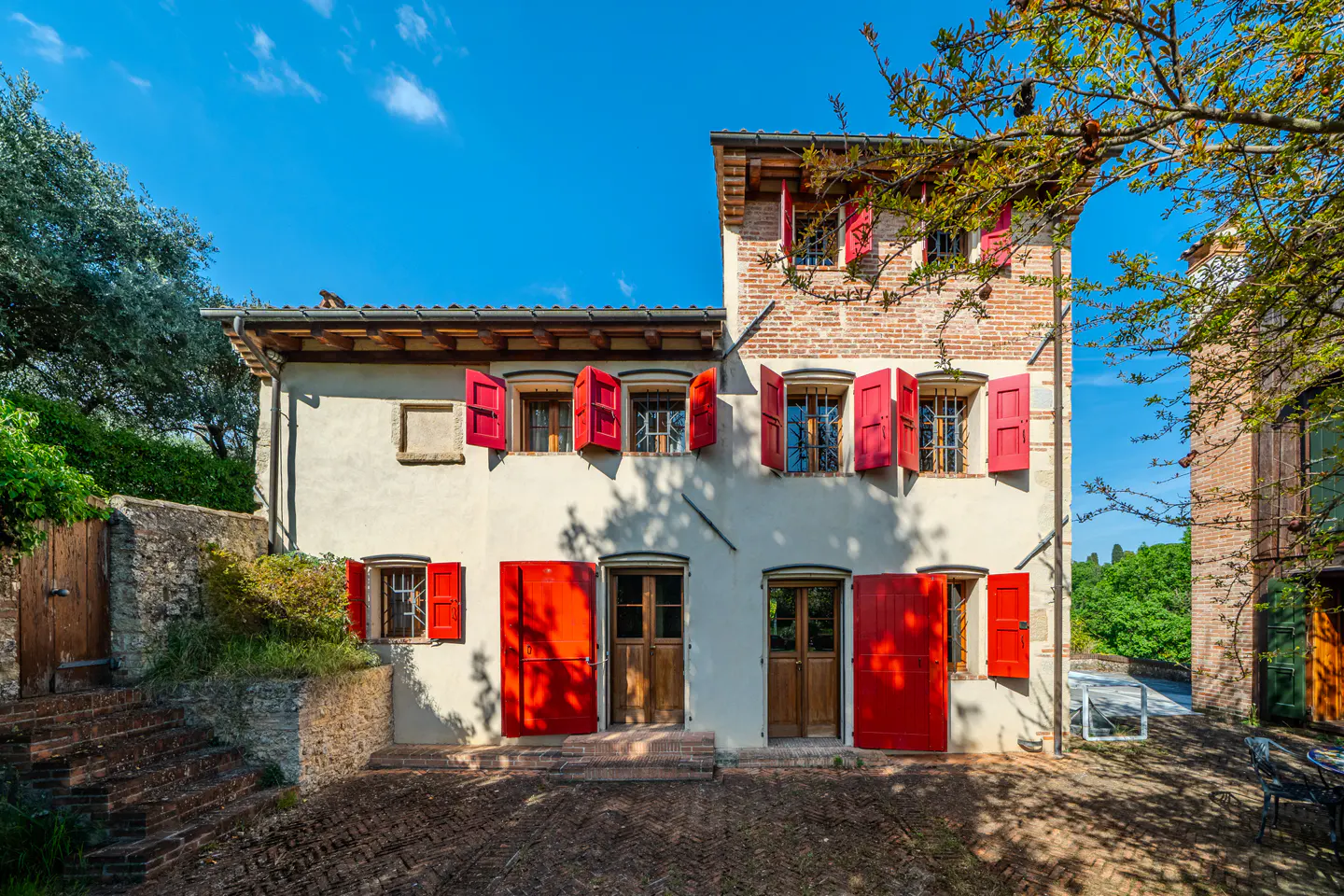 Exterior view of a two-story building with white walls and red shutters under a blue sky. Brick pathway leads to wooden doors.