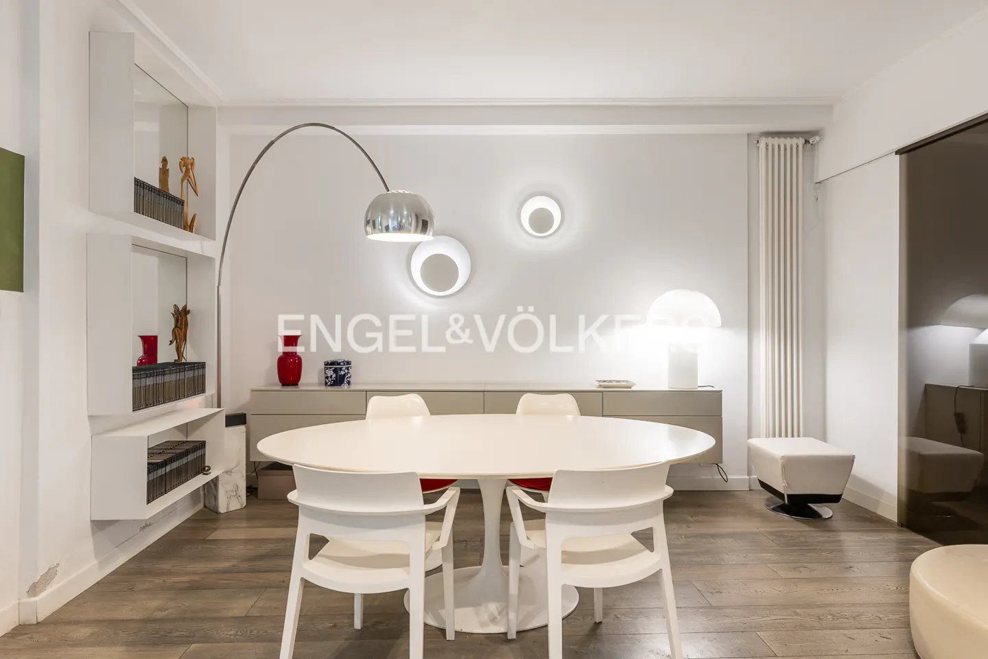 Bright dining room with white oval table, chairs, and modern lighting. A bookshelf and sideboard add storage. Wood floors and neutral tones create a clean look.