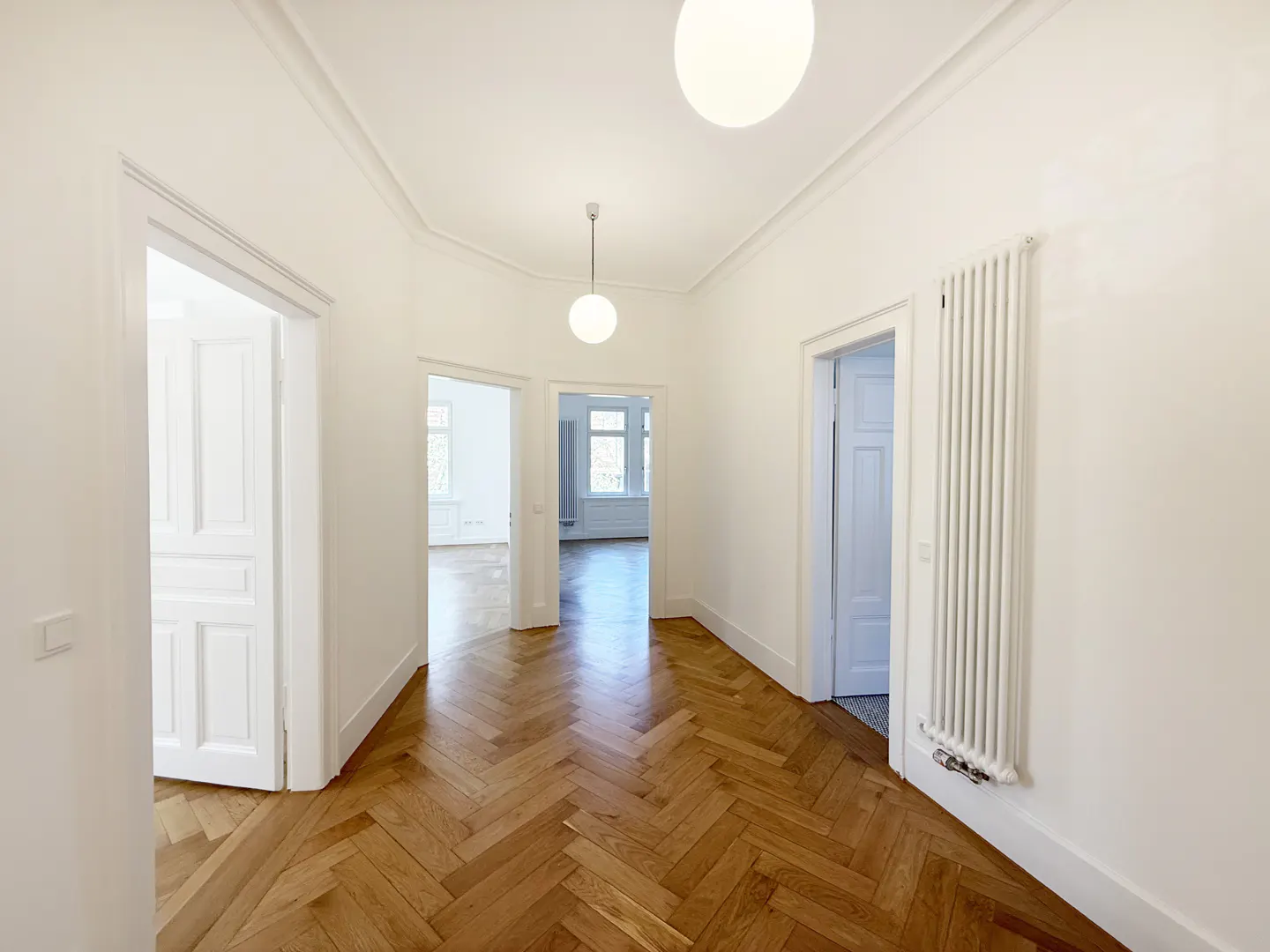 Hallway with herringbone wood floors, white walls, and open doorways. Globe lights hang from the ceiling.