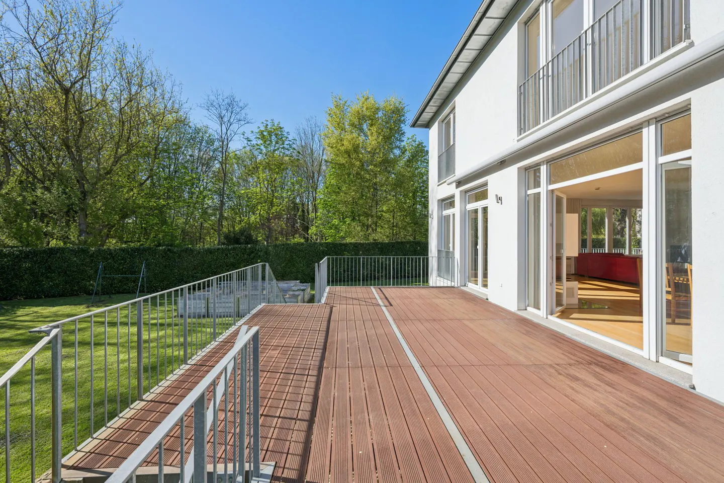 Exterior view of a modern white house with a brown wooden deck, metal railings, and a green lawn with trees. Blue sky.