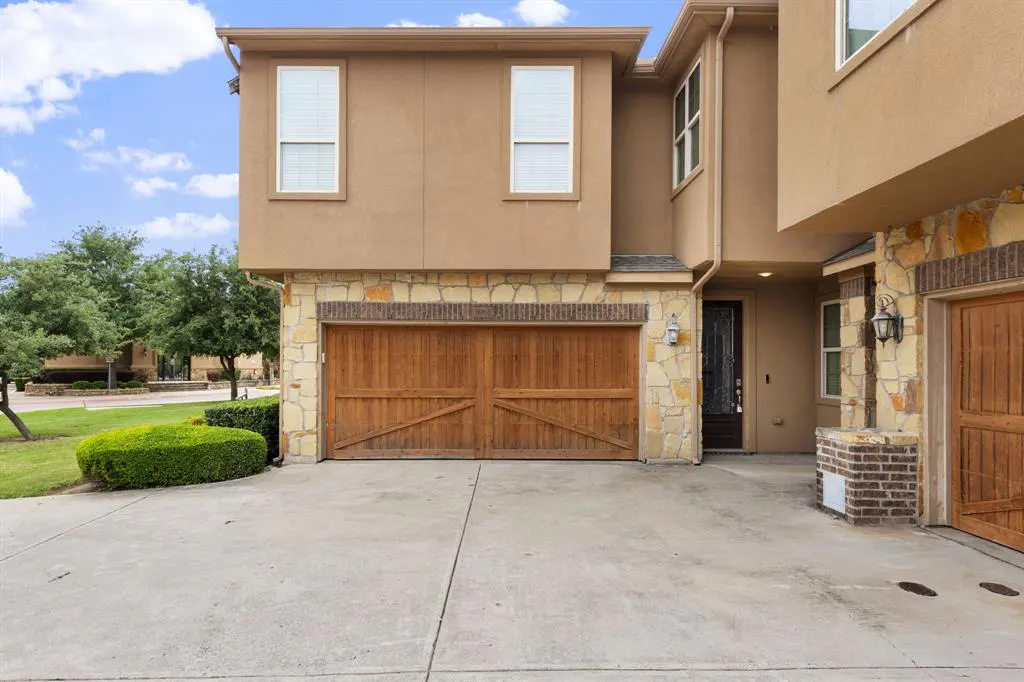 Exterior view of a two-story tan house with stone accents and a wooden double garage door. Windows are visible on both floors.