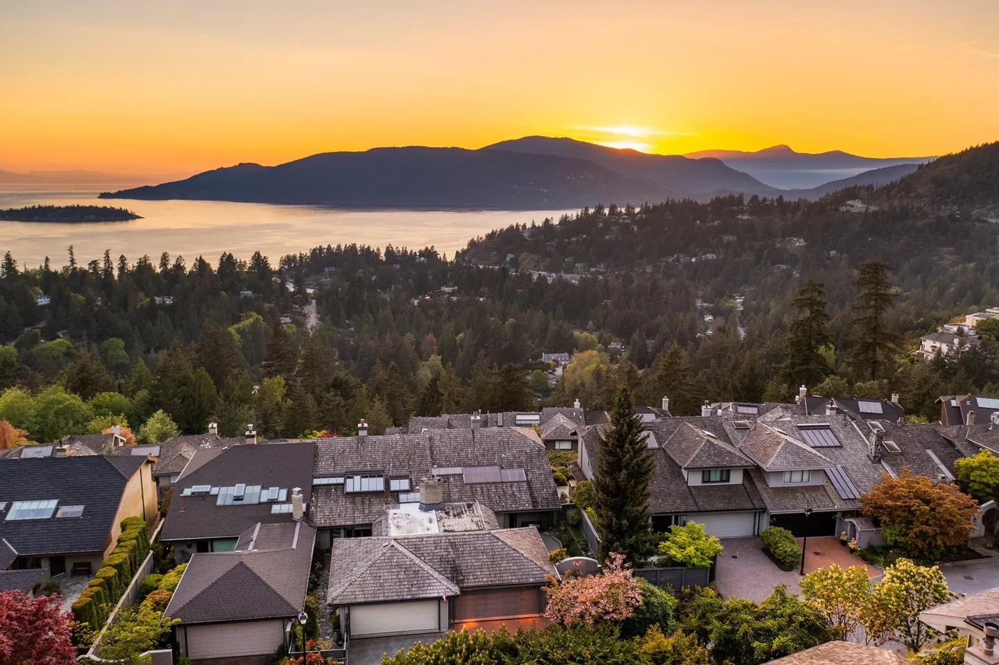 Sunset view of homes nestled in trees, overlooking water and mountains. Orange sky.