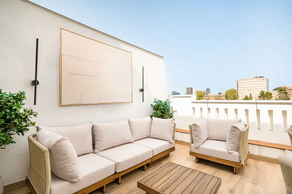 Outdoor patio with a beige sofa, chair, and wood table. A large abstract painting hangs on the white wall. City buildings are visible in the background.