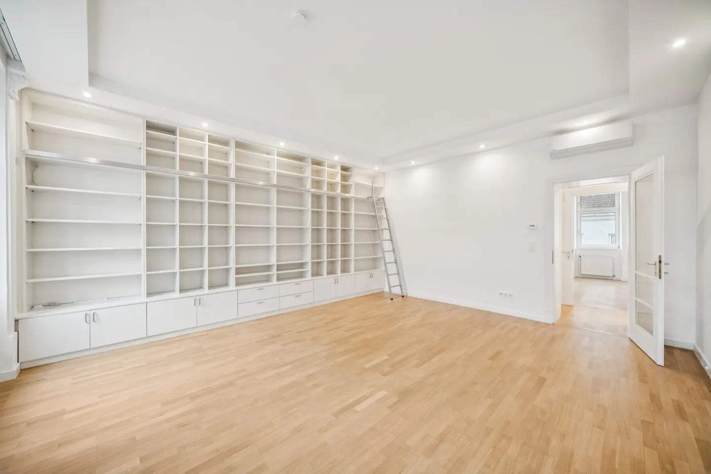 Bright room with light wood floors, a large white wall-to-wall bookshelf with a rolling ladder, and an open doorway.