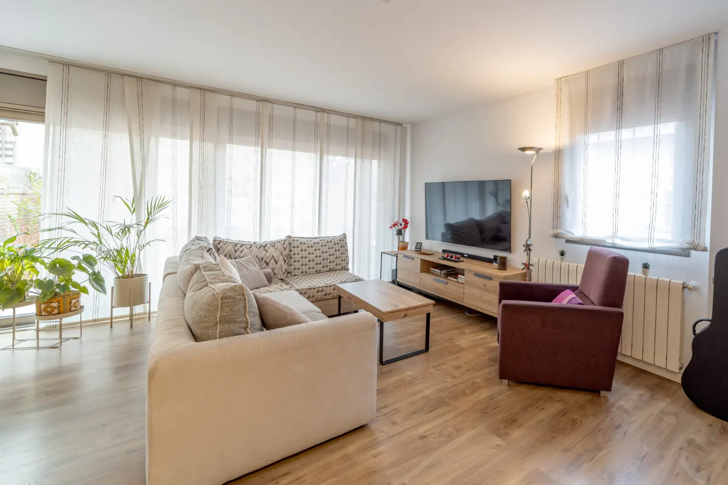 Bright living room with a beige sectional sofa, wood coffee table, TV, and a maroon armchair on a wood floor. Large windows with sheer curtains.