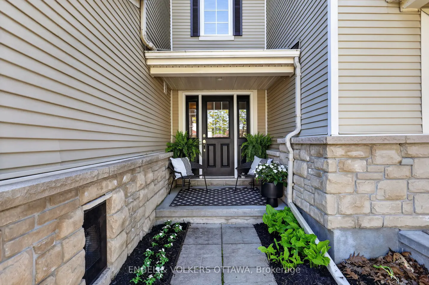 Home's front entrance with a black door, sidelights, and a covered porch. Two chairs with striped pillows flank the door. Ferns and flowers add greenery.
